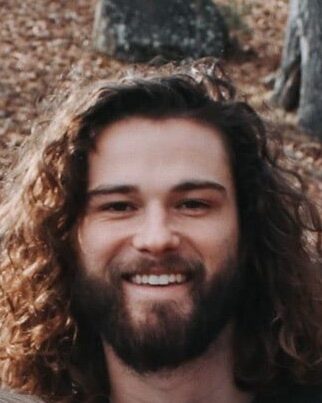 A man with long curly brown hair and a beard is smiling outdoors in a wooded area with fallen leaves and large rocks in the background.