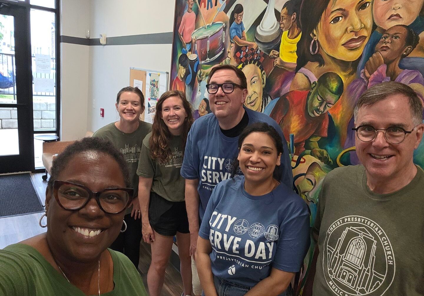 A group of six smiling people pose indoors in front of a colorful mural depicting diverse faces and scenes. Four wear “City Serve Day” shirts, and one person takes a selfie, capturing everyone in the frame.