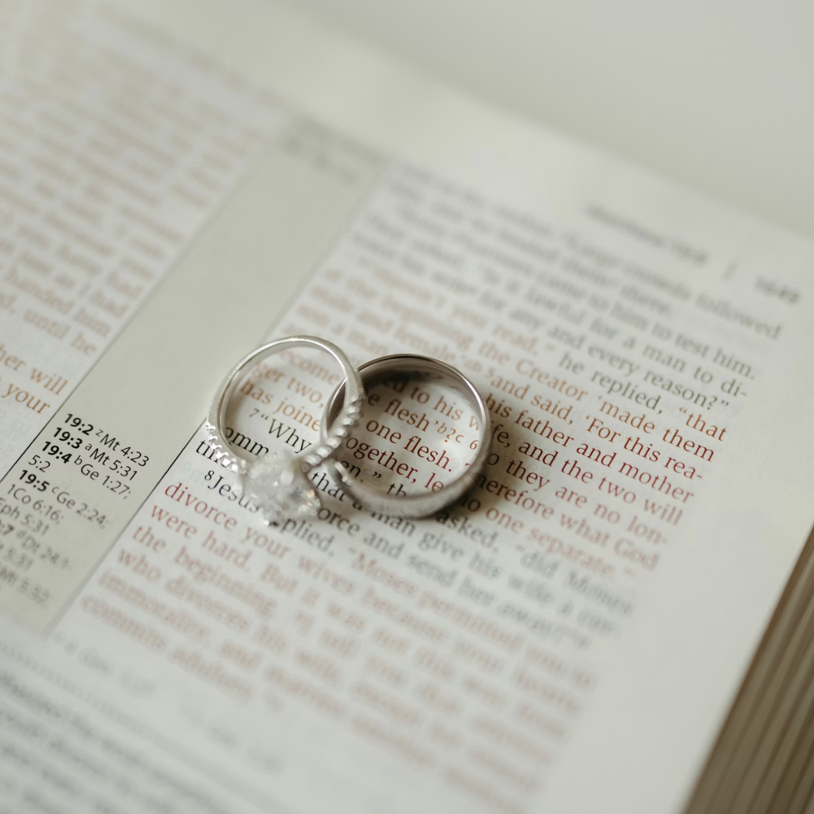 Two rings, one with a diamond and one plain, rest on an open Bible highlighting a passage about marriage. The rings cast a heart-shaped shadow on the page.