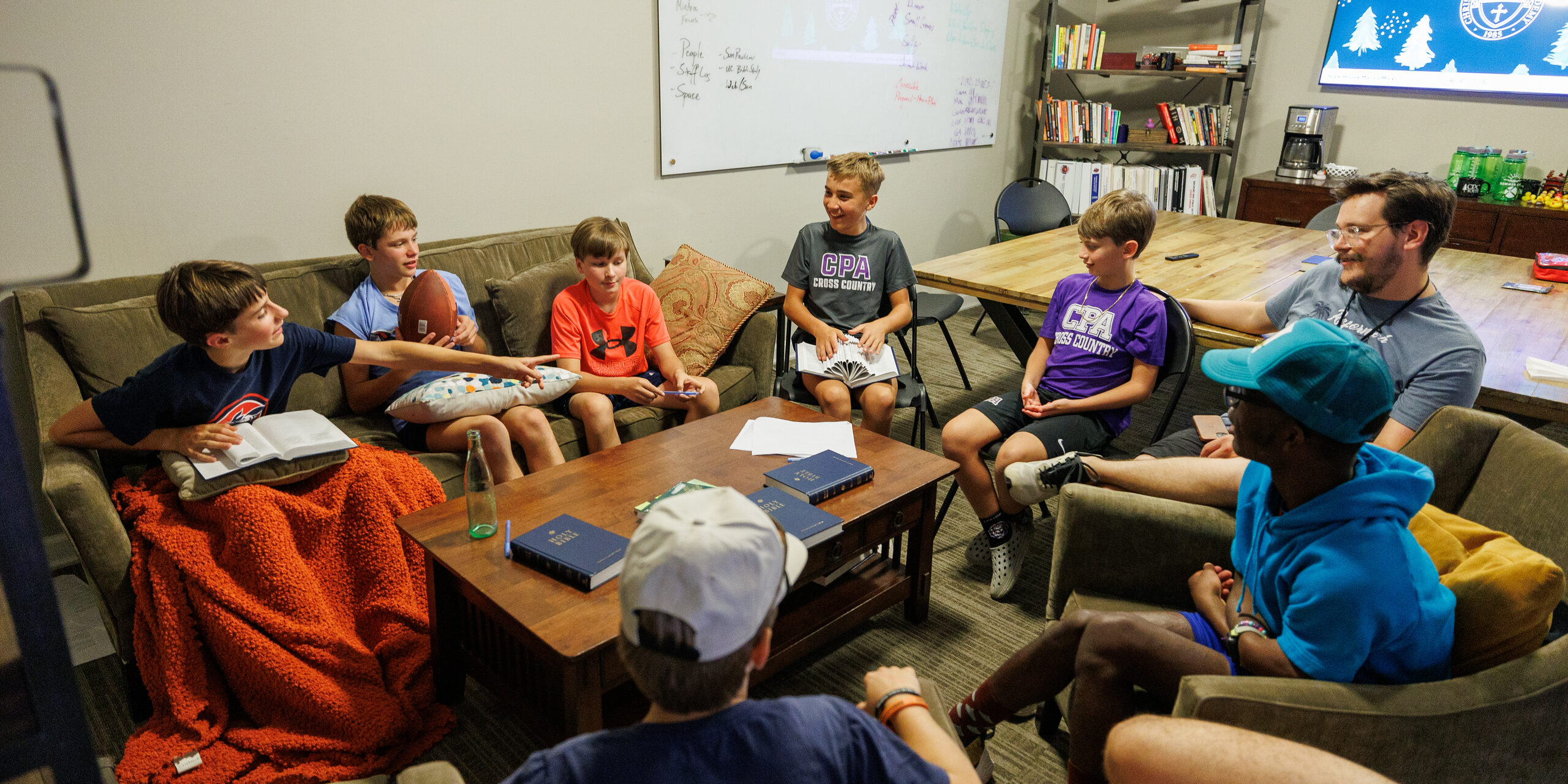 A group of children and one adult sit in a cozy living room, gathered in a circle on sofas and chairs, engaging in discussion. Papers and books are on the table, and a whiteboard and TV screen are in the background.