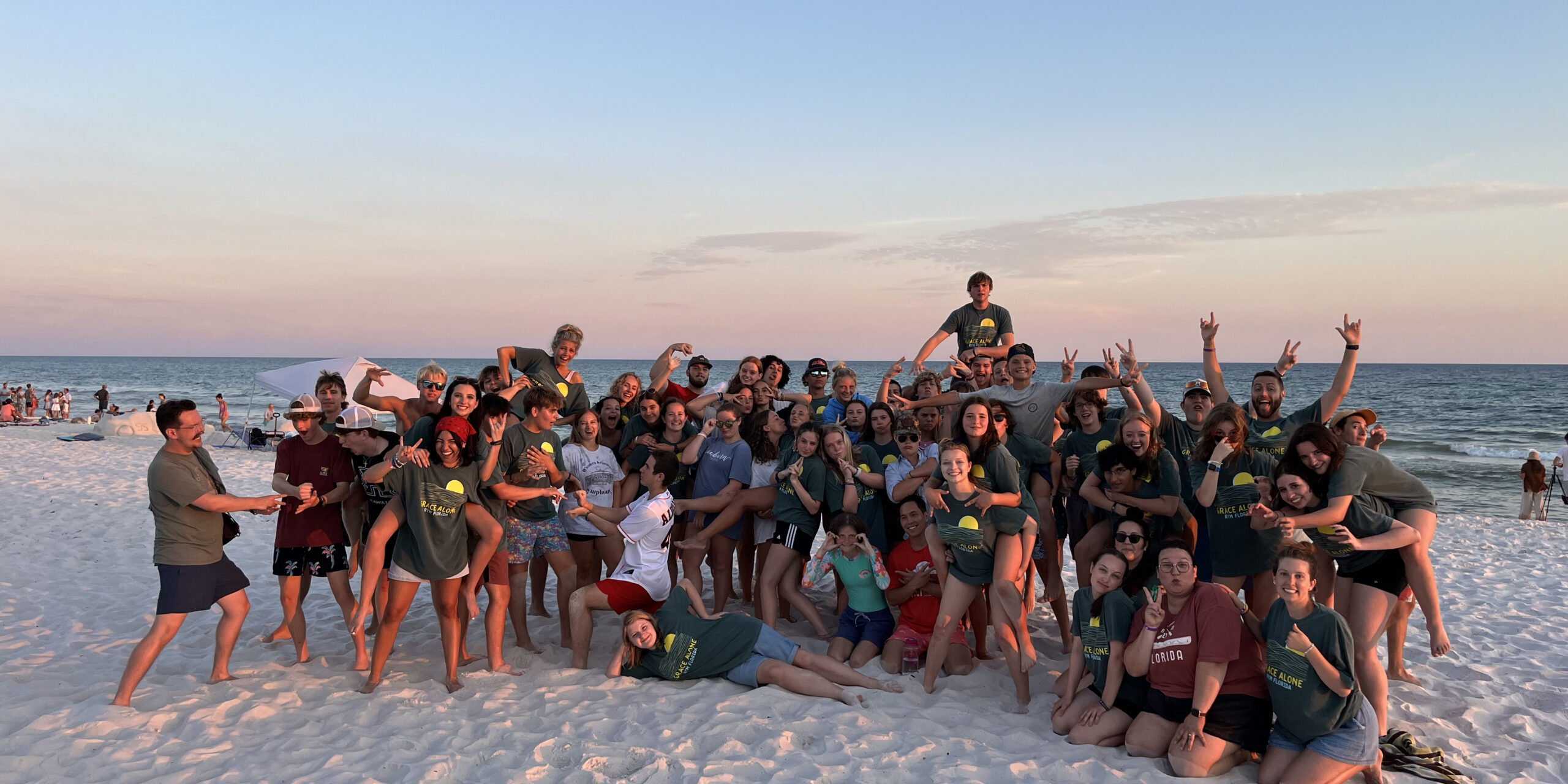 A large group of people, mostly teens, pose playfully on a sandy beach at sunset, wearing matching green shirts. The ocean and sky are in the background, with a few other beachgoers visible in the distance.