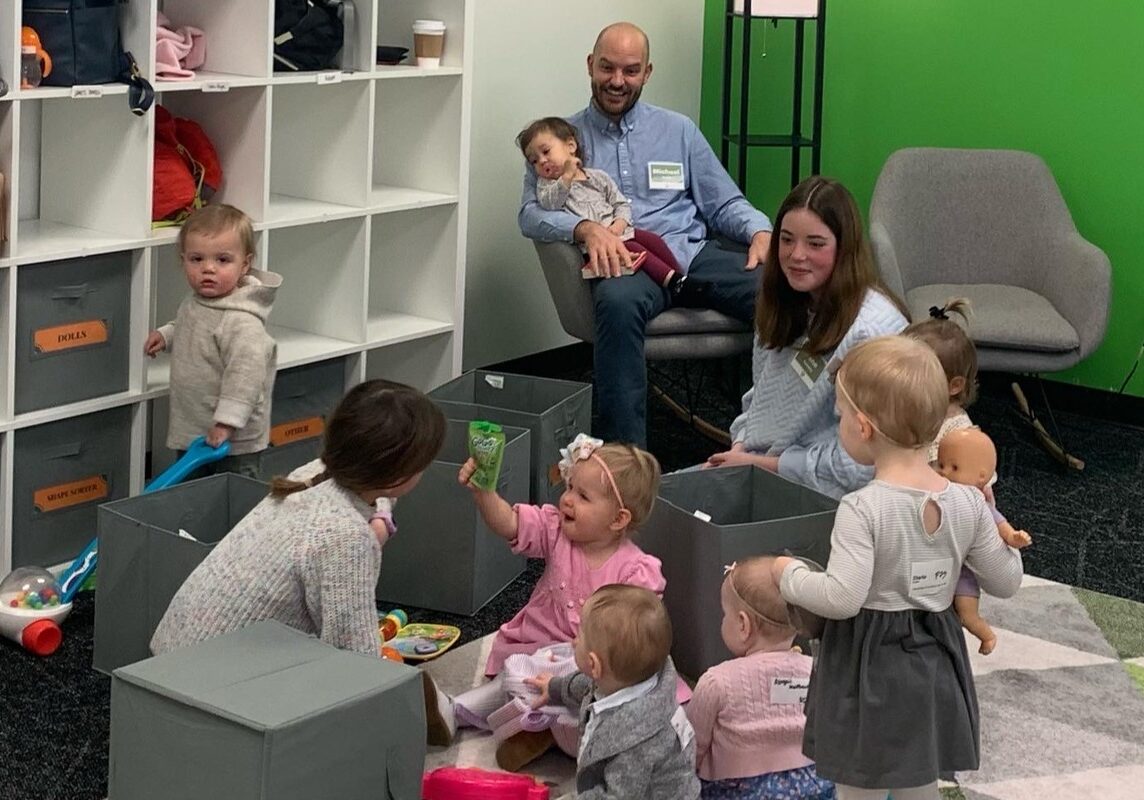 Several young children and two adults are in a playroom. Some kids are playing with toys and dolls on the floor, while an adult sits and another kneels among them. White cubby shelves and a green wall are in the background.