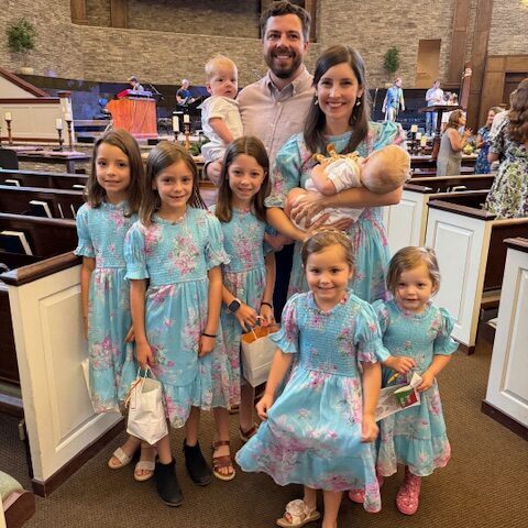 A family poses in a church; six young girls wear matching blue floral dresses, while a woman holds a baby and stands next to a man holding a toddler. Other people are in the background near pews and an altar.