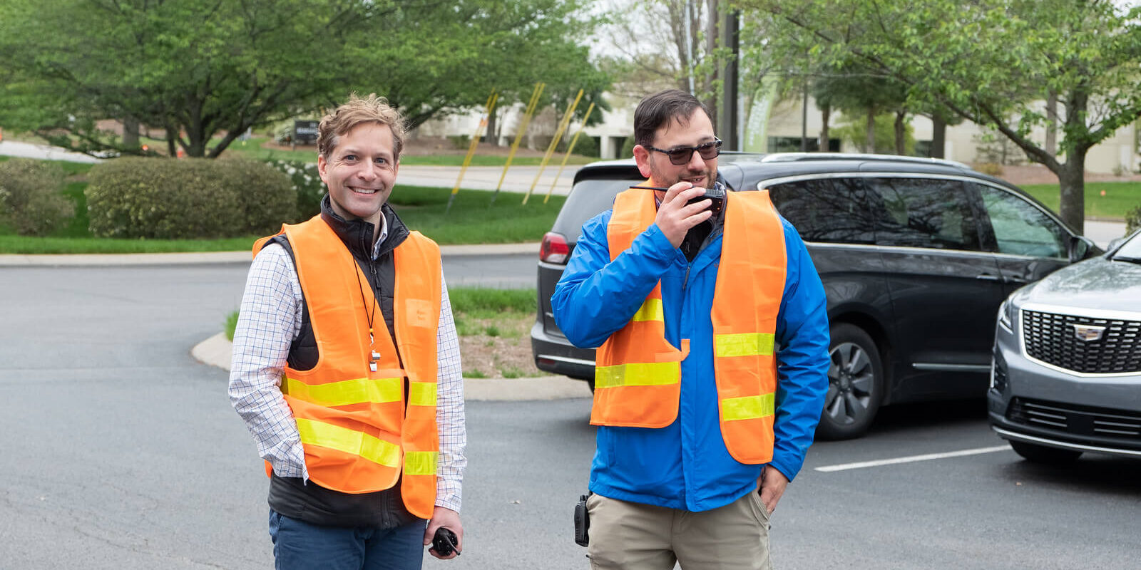 Two men wearing orange safety vests stand in a parking lot. One is smiling with hands in his pockets, while the other speaks into a walkie-talkie. Trees and parked cars are visible in the background.