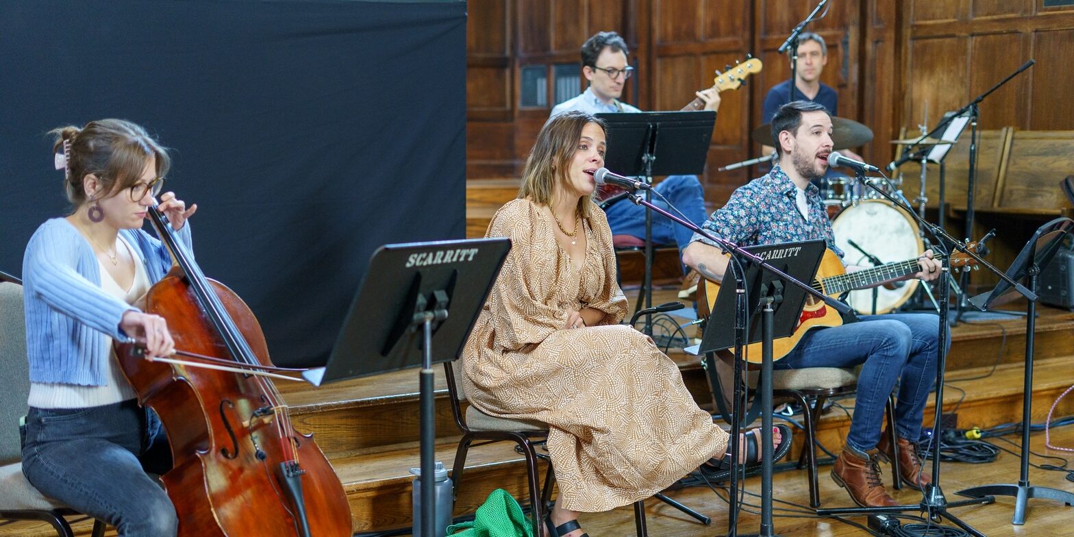 A band performs indoors; a woman plays cello, a woman sings into a microphone, a man sings and plays acoustic guitar, while two men in the background play bass and drums. Music stands and wooden walls are visible.