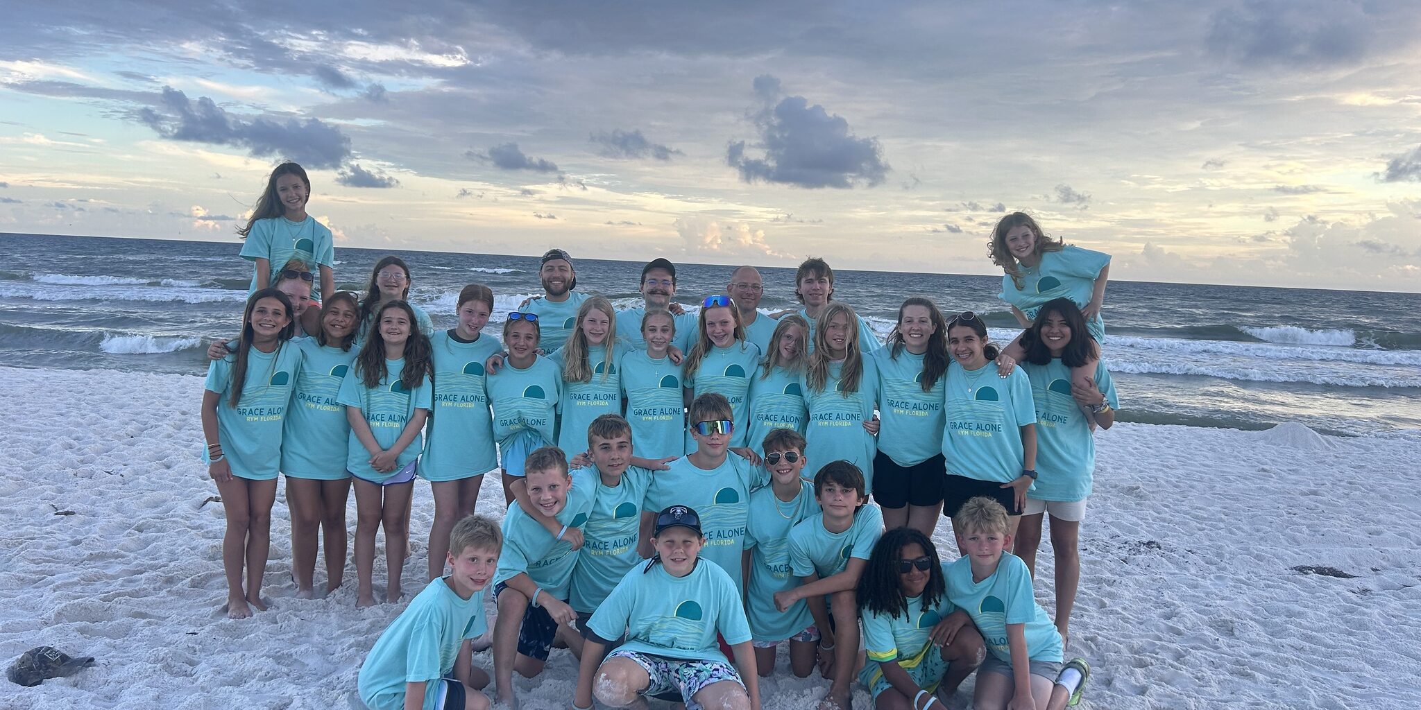 A group of children and adults in matching teal shirts pose together on a sandy beach with the ocean and a cloudy sky in the background.